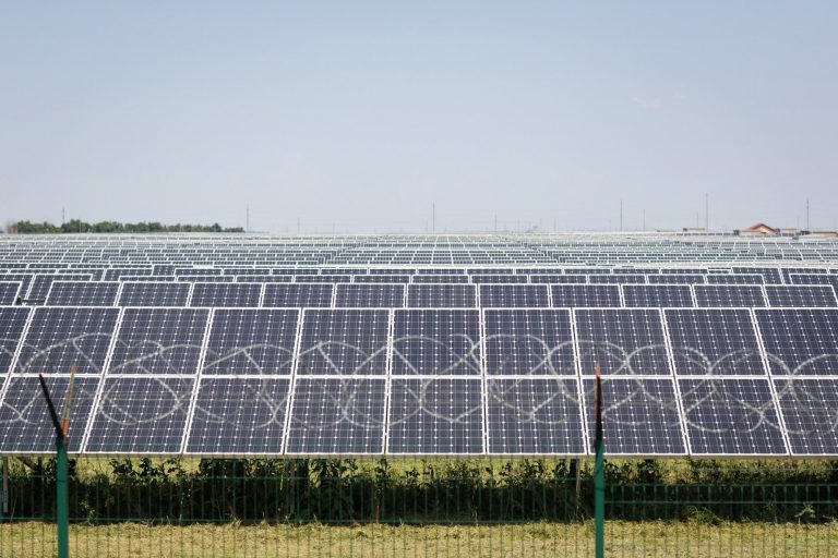 Bild einer industriellen Großanlage zur Erzeugung von Solarstrom. Im Vordergrund ein hoher Zaun mit Stacheldrahtkrone.